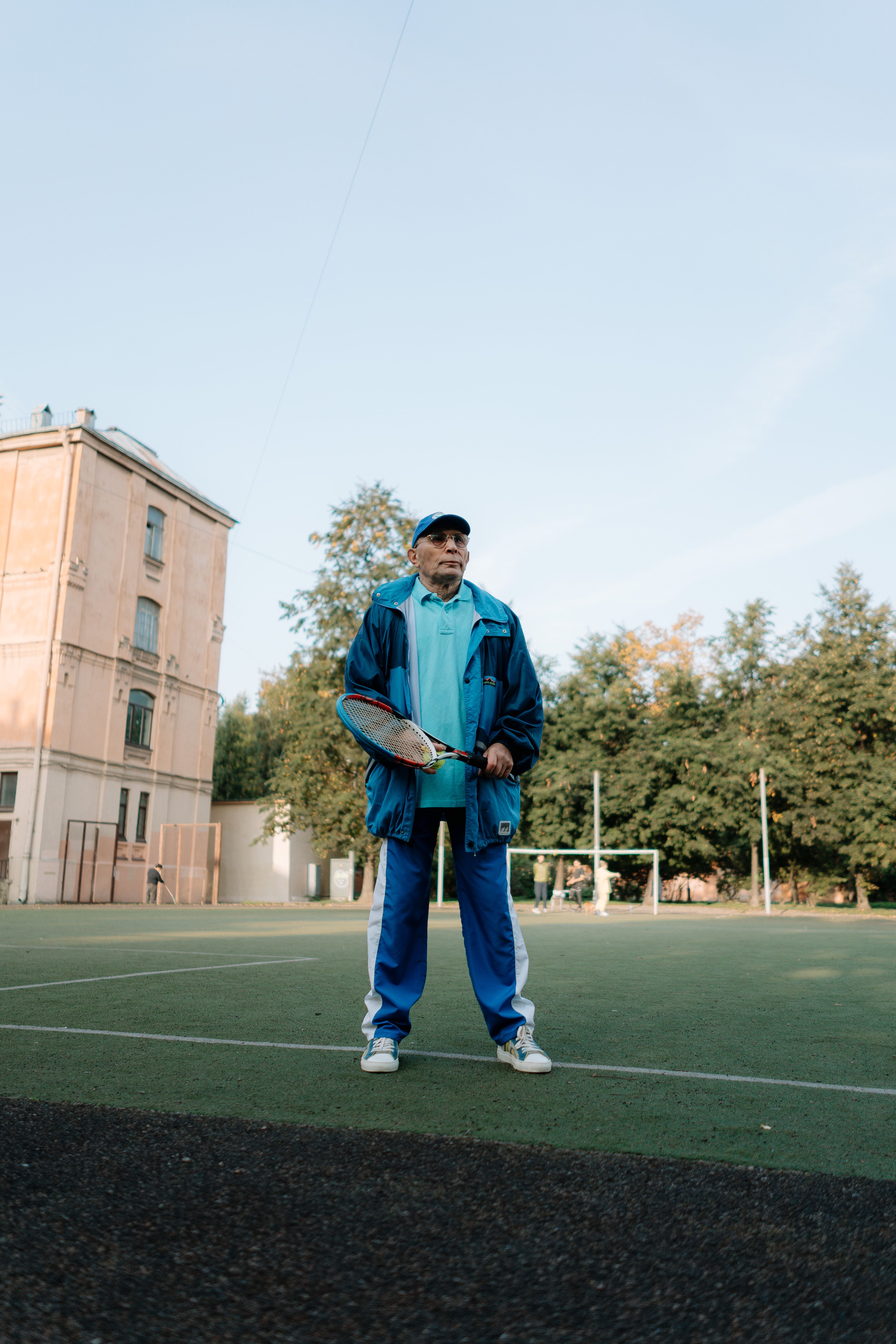 Elderly man in blue sports attire holding a tennis racket on an outdoor court.