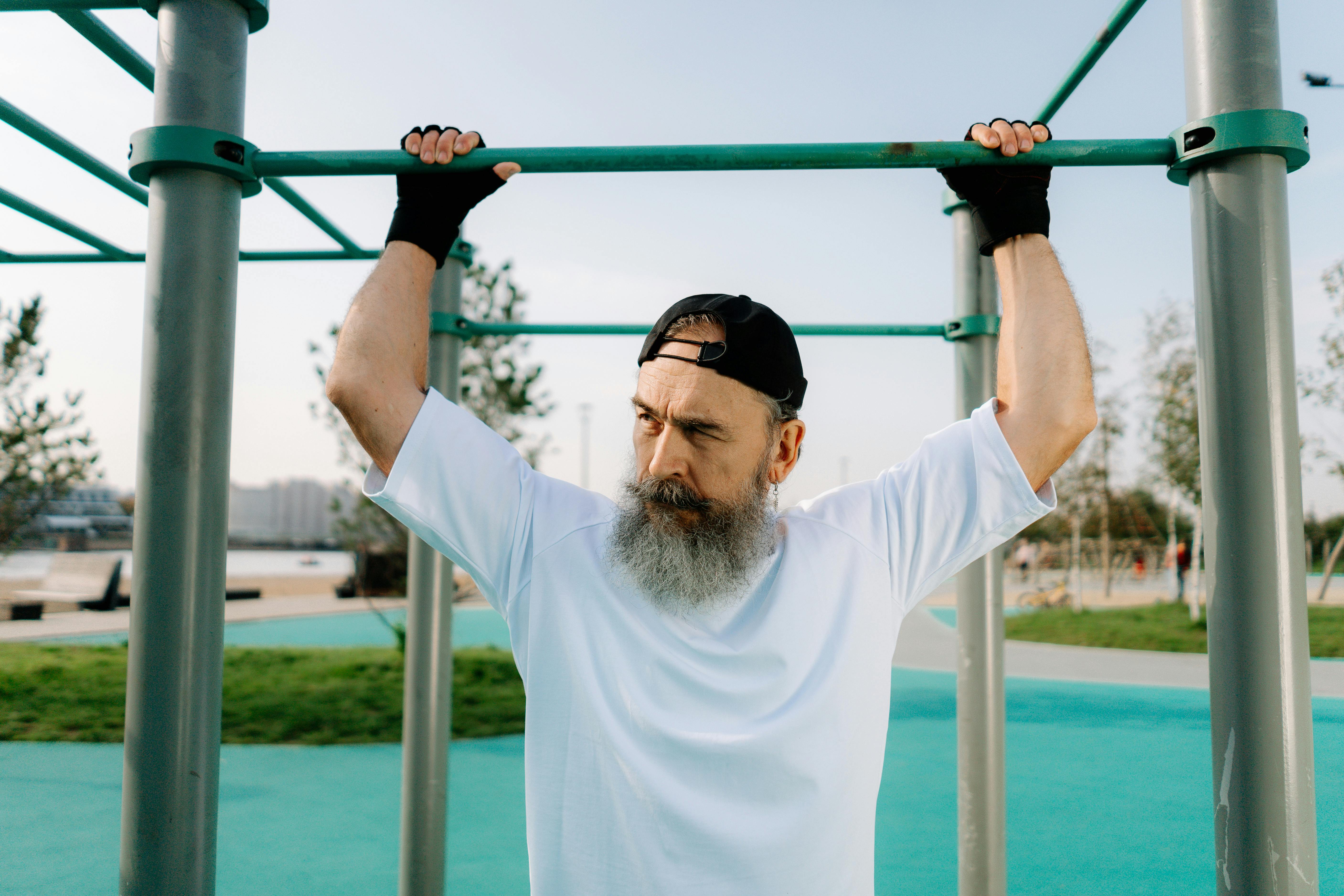 bearded man in white crew neck shirt and black cap holding on to a monkey bar