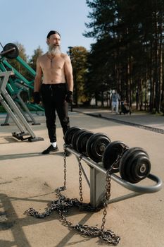 A shirtless senior man exercises outdoors with dumbbells on a sunny day.