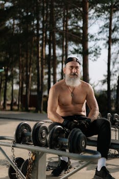 Shirtless bearded man sitting outdoors, surrounded by dumbbells, in a fitness setting.