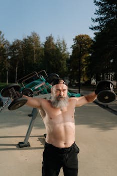 A shirtless, bearded man lifting dumbbells in an outdoor fitness area.