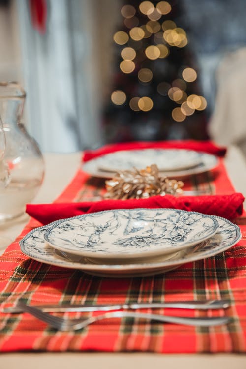 Table with silverware and porcelain plates placed on checkered red napkins near jug with  blurred garland on Christmas tree on background