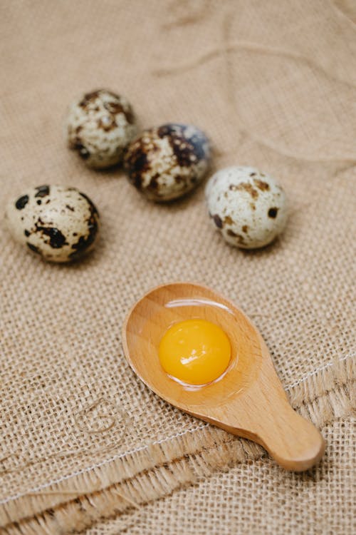 From above of timber spoon with fresh yolk near spotty quail eggs on canvas fabric