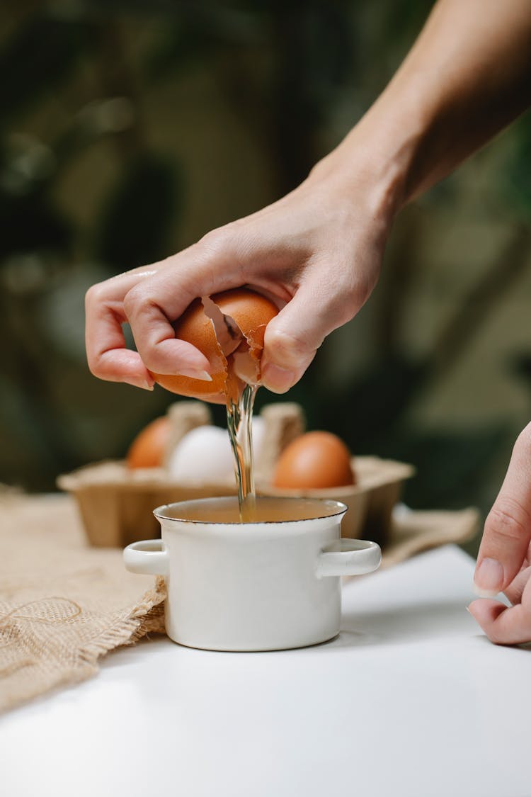 Woman Breaking Chicken Egg In Saucepan On Table