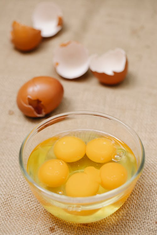 High angle of glass bowl with raw eggs placed near eggshell for cooking process