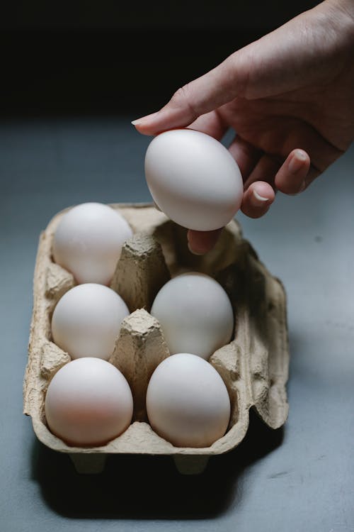 High angle of crop anonymous female taking raw chicken egg from cardboard container for cooking