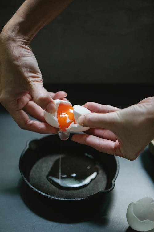 High angle of crop anonymous man breaking egg on frying pan for making delicious breakfast in kitchen