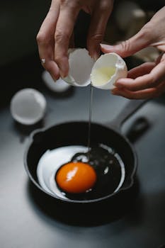 High angle of crop anonymous lady breaking egg in metal frying pan during preparation of breakfast in kitchen