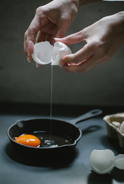 Free Crop anonymous female cook breaking white chicken egg into small iron frying pan placed on table in kitchen Stock Photo