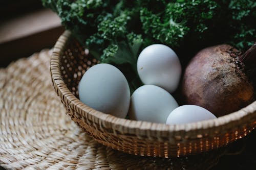 From above of fresh raw eggs with beetroot and bunch of green herbs placed in wicker basket on table