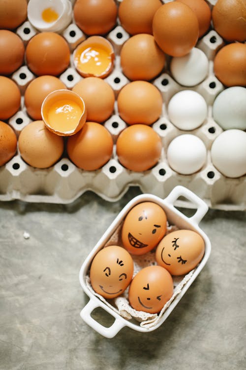 Free From above of square bowl with brown chicken eggs with painted faces placed near carton box with assorted production in kitchen Stock Photo
