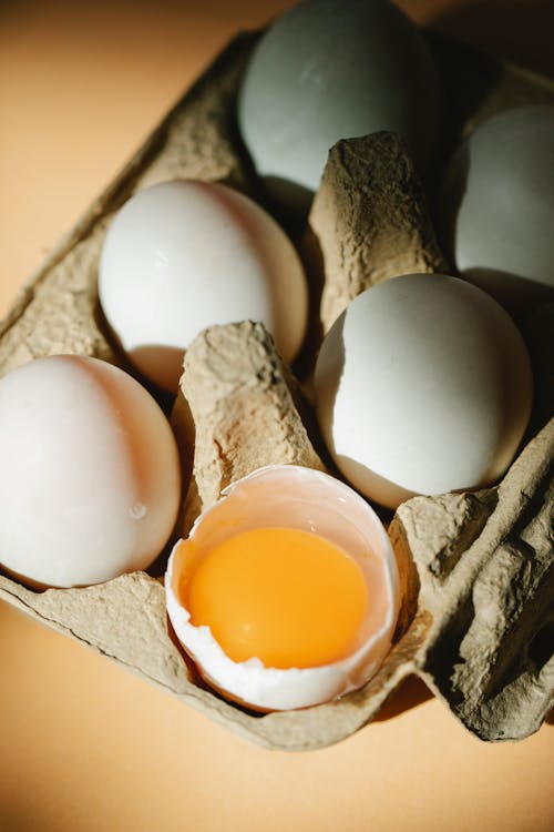 From above of whole and broken white raw chicken eggs with bright yolk placed in carton container