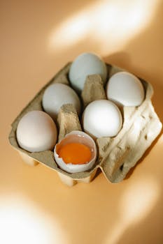 High angle of carton container with fresh organic whole and broken eggs with bright yolk placed on beige table in daylight