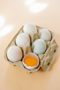 High angle of broken white chicken egg with vibrant yolk placed in filled carton box on beige surface in kitchen in sunny morning