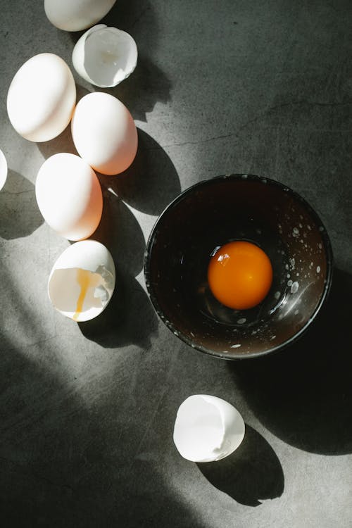 To view composition of raw fresh cracked egg in bowl placed on table near scattered white eggs and eggshells
