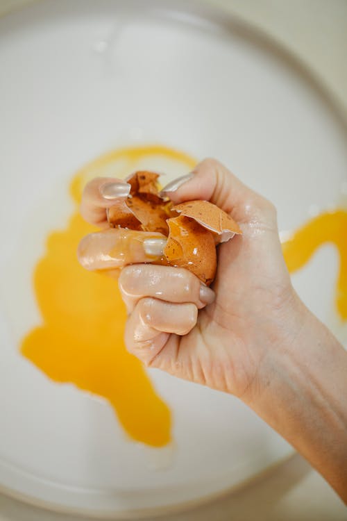 Free From above crop anonymous female breaking raw brown egg with manicured hand above white plate in kitchen Stock Photo