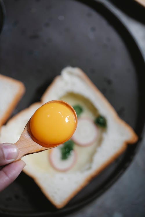 Free Top view crop anonymous chef demonstrating yolk in spoon while cooking delicious egg toast on pan for breakfast Stock Photo