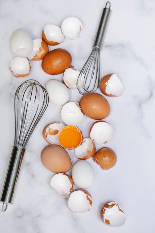 Overhead arrangement of broken eggshells and whole raw brown eggs placed on kitchen marble table near whisks