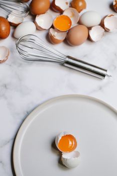 Top view of broken egg with yolk on white round plate placed on table with heap of eggshells and whisk in kitchen