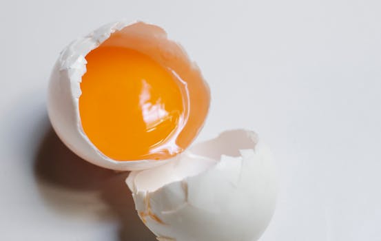 Top view of broken raw egg with yellow yolk and white eggshell on white background in light kitchen during cooking process