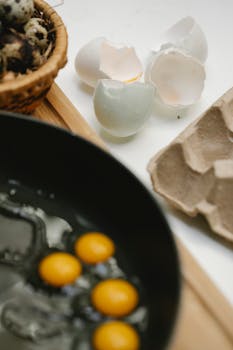 From above of small quail eggs frying in pan near eggshells and egg carton on white table in light kitchen
