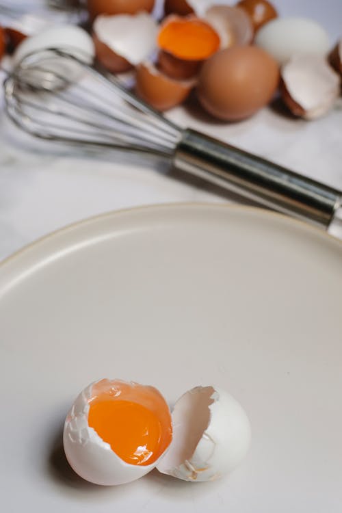 Free From above of raw broken egg on round plate placed on table with pile of eggshells and whisk on blurred background Stock Photo