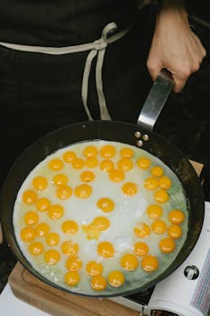 High angle crop anonymous chef in black apron frying delicious raw quail eggs on pan in kitchen