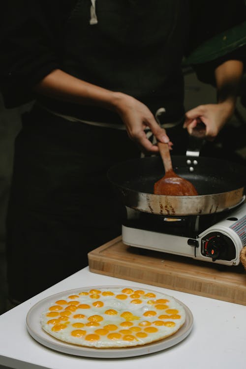 Crop anonymous cook placing wooden spatula in pan placed on stove near freshly fried quail eggs on plate