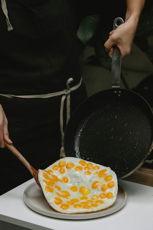 Crop anonymous cook with spatula putting freshly fried quail eggs from pan on plate