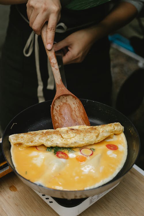 High angle crop anonymous chef rolling yummy omelette with tomatoes and radish on frying pan by using wooden spatula