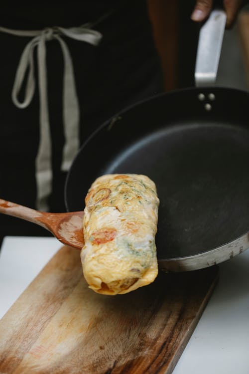 Crop anonymous chef in apron putting fresh omelette roll from frying pan on wooden board in kitchen