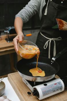 Crop unrecognizable cook wearing black apron pouring beaten eggs with vegetables on hot frying pan in kitchen