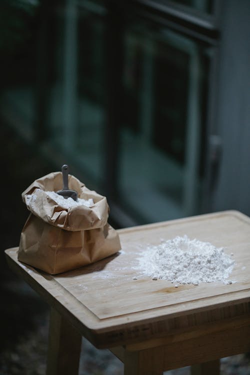 Composition of baking flour scattered on wooden table near flour craft bag with shovel in dark kitchen