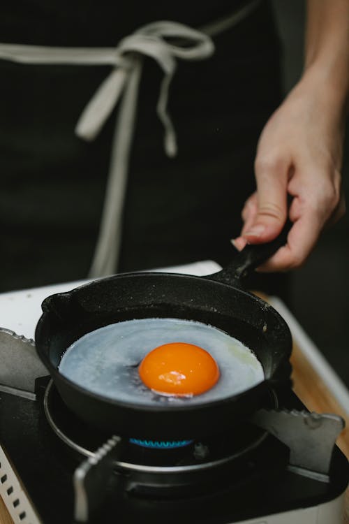 Free Crop anonymous housewife wearing black apron frying raw chicken egg on small pan in kitchen Stock Photo