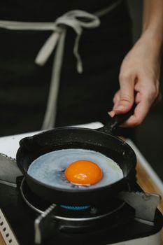 Crop anonymous housewife wearing black apron frying raw chicken egg on small pan in kitchen