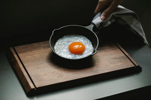 Crop anonymous chef demonstrating yummy fried chicken egg on pan placed on wooden board in dark kitchen