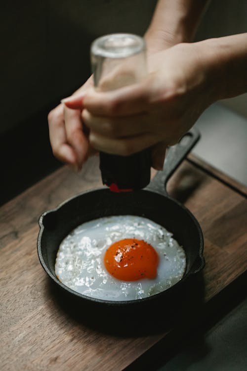 From above of crop anonymous female pouring savory from pepper mill on portion of fried egg in pan