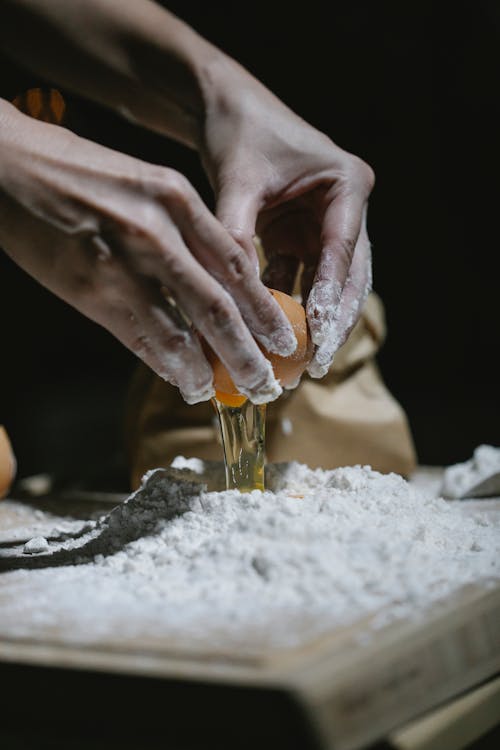 Crop anonymous baker breaking egg to flour while standing at table and cooking dough