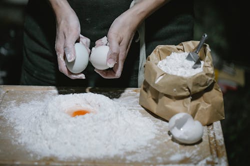 High angle of crop anonymous chef breaking eggs for cooking dough at table with flour