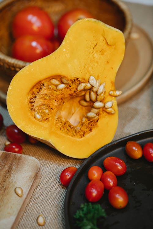 From above of ripe pumpkin with seeds placed near bowl of tomatoes on table for cooking