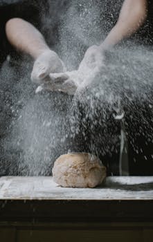 Crop anonymous chef standing at table and scattering flour on dough during cooking process