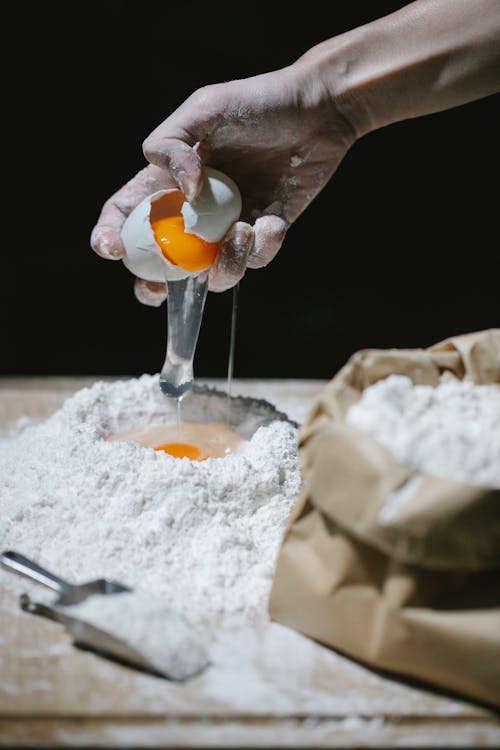 Crop unrecognizable cook adding egg in pile of flour for preparing dough at wooden table