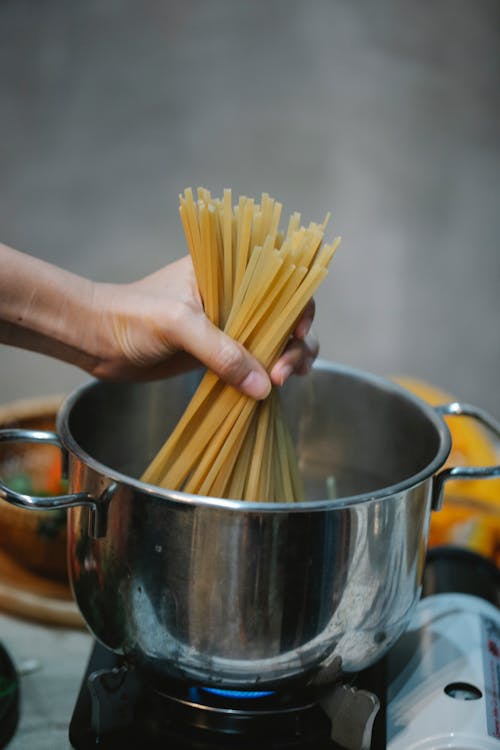 High angle of crop anonymous female putting uncooked pasta into saucepan heating on gas stove in kitchen