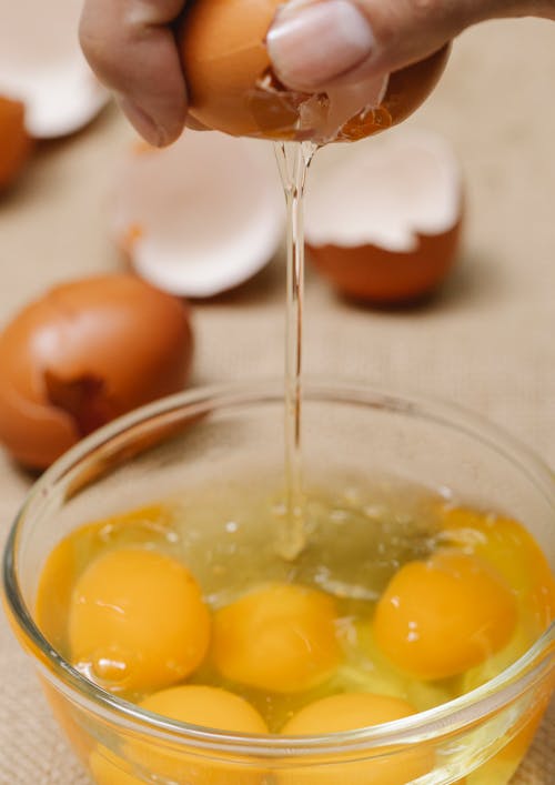 High angle of crop anonymous female adding egg into bowl while cooking in kitchen