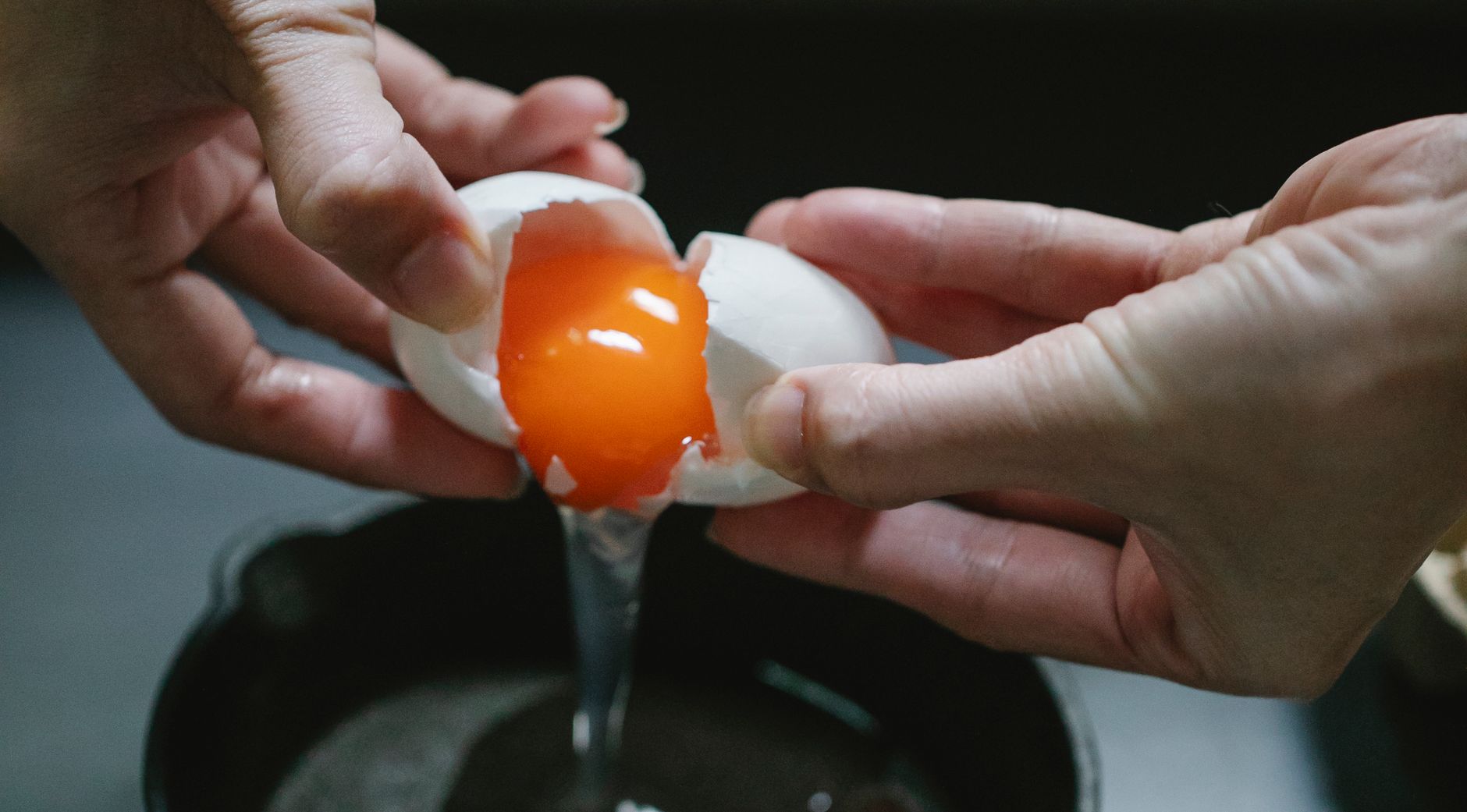 Crop anonymous chef breaking raw chicken egg in pan against dark blurred background