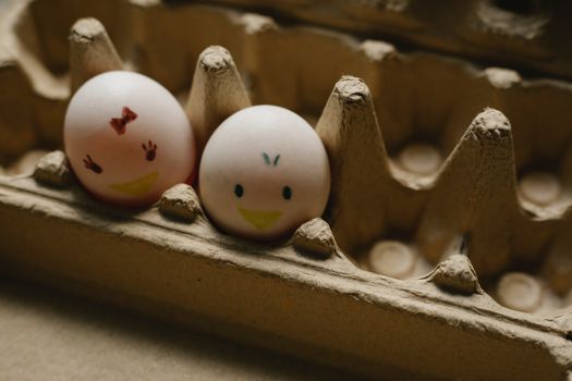 From above of white chicken eggs with funny faces placed in recycled carton box on table