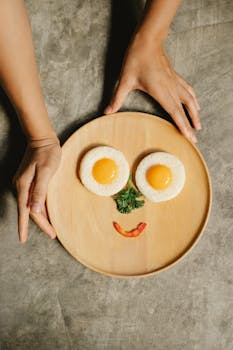 A playful breakfast arrangement featuring fried eggs and vegetables forming a smiley face on a wooden plate.