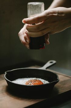 Crop anonymous female chef milling pepper in frying egg in pan placed on wooden board against blurred background