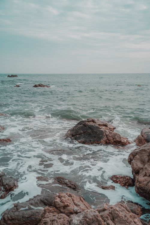 Free A serene seascape with waves crashing on a rocky shoreline under a cloudy sky. Stock Photo