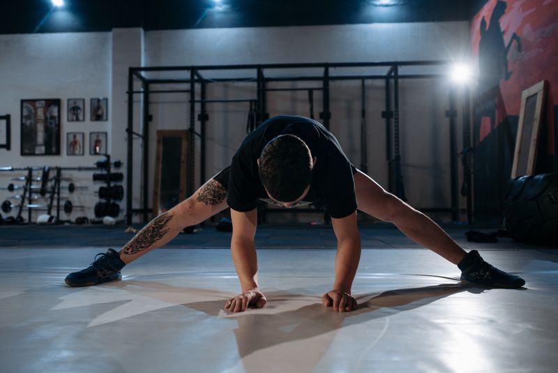 A man with a tattoo stretches on a gym floor under dramatic lighting.